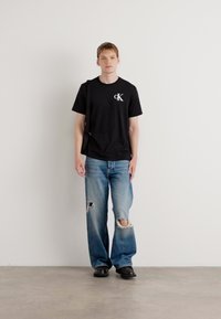 Black cotton t-shirt with CK logo, light blue ripped jeans, black shoes, and a black crossbody bag, standing against a white background.