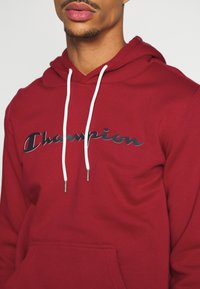 Sweat à capuche rouge avec des cordons blancs et une poche kangourou à l'avant. Présente un logo "Champion" noir en cursive sur la poitrine.