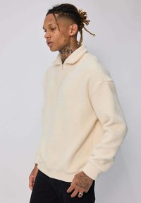 Pull en polaire crème avec un col zippé, coupe décontractée et poignets côtelés. Présente une texture douce et des coutures minimales pour un look épuré.