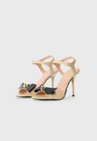Sandaile cu toc înalt au un superior din piele lacuită nude, accent cu fundiță neagră și detalii aurii, și o brăță la glezna. Design cu vârful deschis.