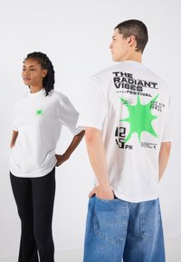 White cotton T-shirts featuring green graphic designs on the front and back. One shirt includes festival text, the other has a star pattern.