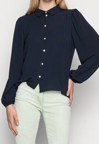Navy blue blouse featuring a collar, long puff sleeves, and eight metallic buttons down the front. Soft, smooth fabric with a relaxed fit.