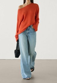 Femme portant un pull orange à épaules dénudées, un jean bleu clair à jambes larges, des sandales noires et portant un sac à main noir tissé, marchant vers l'avant.