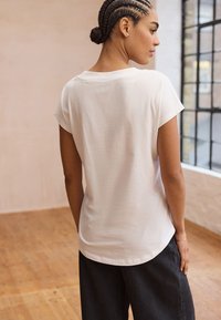 Weißes Baumwoll-T-Shirt mit kurzen Ärmeln, abgerundetem Saum und weicher Textur. Modell von hinten in einem minimalistisch eingerichteten Innenraum gezeigt.