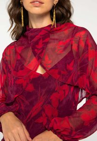 Blouse en tissu transparent à motifs floraux rouges avec superposition, manches longues avec poignets froncés et un design en trou de serrure au niveau du col.