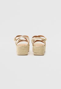 Des sandales dorées à lanières présentent une semelle compensée en espadrille tissée, une surface texturée et une fermeture à boucle réglable. La vue arrière met en valeur le design et la hauteur.