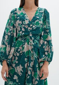 Robe à imprimé floral en bleu sarcelle et beige avec un col en V, des manches bouffantes et une taille froncée. Fabriquée en tissu léger et texturé.