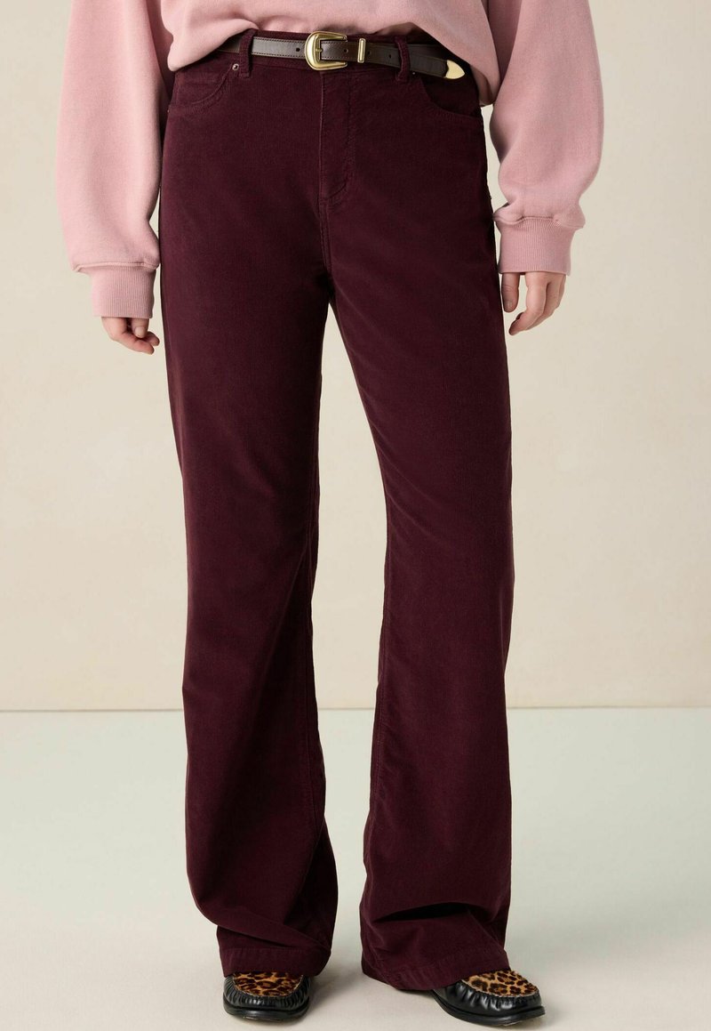 Personne portant un pantalon en velours côtelé bordeaux foncé avec une ceinture noire, un haut à manches longues rose et des chaussures noires avec des orteils en imprimé léopard.