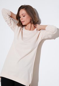 Top de manga larga beige claro con cuello redondo, puños acanalados, aberturas laterales y un corte relajado, hecho de suave material de algodón.