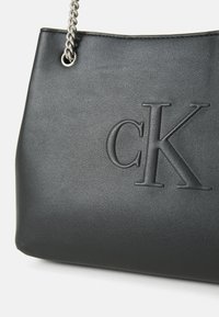 Čierna syntetická kabelka s embosovaným logom "CK", s hladkou textúrou a strieborným reťazovým popruhom. Dizajn je obdĺžnikový s zaoblenými hranami.