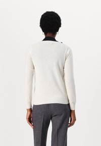 Osoba s krátkými kudrnatými vlasy, oblečená v bílém svetr s dlouhým rukávem, šedých kalhotách a zlatých náušnicích ve tvaru kruhu, zachycena zezadu na bílém pozadí.