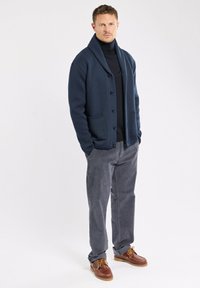 Homme portant un cardigan boutonné bleu marine, un col roulé noir, un pantalon en velours côtelé gris et des chaussures en cuir marron, se tenant avec les mains dans les poches.