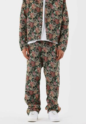 Homme portant une veste et un pantalon fleuris assortis dans des tons rouges, roses, beiges et verts atténués, avec des baskets blanches et une chemise blanche en dessous.