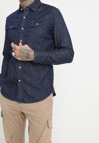 Hombre con la mano y los dedos tatuados, vistiendo una camisa de mezclilla azul oscuro y pantalones cargo beige, de pie contra un fondo blanco liso.