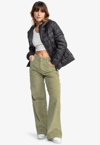 Zwarte pufferjack met gestikte details, een cropped witte tanktop en olijfgroene corduroy wijde broek. Witte sneakers maken de outfit af.