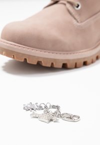 Botte beige clair à semelle épaisse au-dessus d'un bracelet en argent orné d'un charme fleur, un arrosoir avec des strass, et un mousqueton à fermeture.