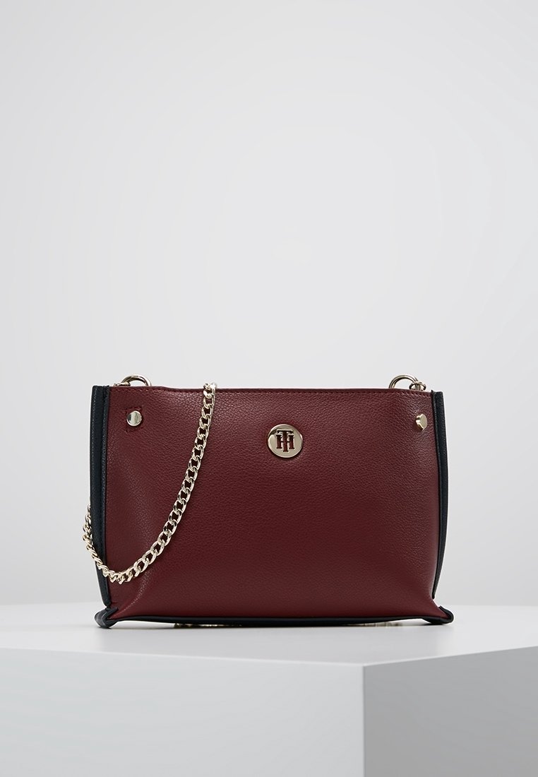 Bolso de cuero burdeos con fondo navy, herrajes dorados, correa de cadena y detalle del logo en la parte delantera. Textura suave, forma estructurada.