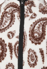 Vesta z fleecu s bílým podkladem a hnědými paisley vzory, černým předním zipem a měkkým, texturovaným povrchem.