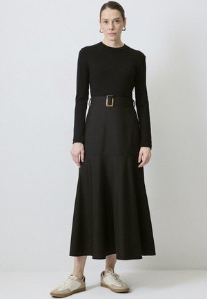 Žena stojaca v čiernych šatách s dlhým rukávom, širokým opaskom a béžovými teniskami na svetlom jednoliatom pozadí.