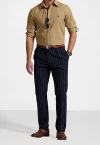 Beżowa koszula na guziki, granatowe spodnie w prążki, brązowy skórzany pasek i buty. Czarne okulary przeciwsłoneczne podkreślają elegancki wygląd stroju.