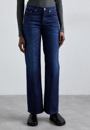 Jeans a gamba larga blu scuro in denim, con un classico design a cinque tasche e accessori in metallo, abbinati a calzature nere.