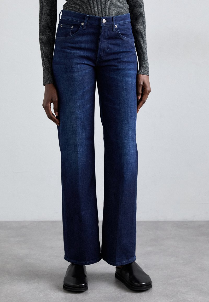 Jeans de pierna ancha de color azul oscuro, hechos de mezclilla, que cuentan con un diseño clásico de cinco bolsillos y herrajes metálicos, combinados con calzado negro.