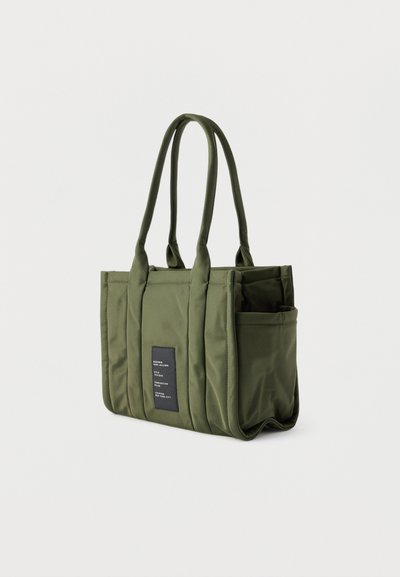 Τσάντα ώμου από ύφασμα σε ελιά πράσινο, με διπλούς ιμάντες ώμου, πλαϊνή τσέπη και μαύρη ορθογώνια ετικέτα με λευκό κείμενο στο μπροστινό μέρος.