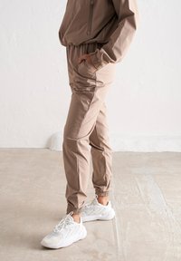 Joggers beiges en tissu lisse avec une taille élastique et des poignets élastiques. Comprend des poches latérales et un logo discret sur la cuisse. Associés à des chaussures blanches.