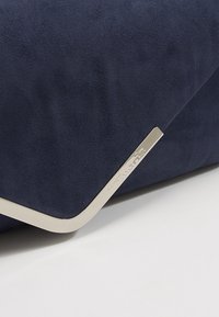 Gros plan sur un coin de sac en daim bleu marine avec un bord métallique argenté gravé du nom de la marque, sur fond blanc.