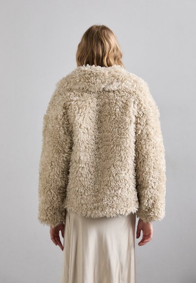 Beige faux fur jas met een rond kraag, oversized pasvorm en een gestructureerd, harig oppervlak. De achterkant toont een naadloos ontwerp en omgeslagen mouwen.