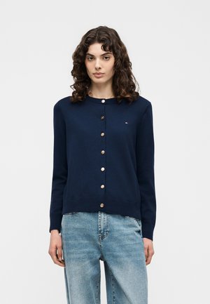 Vrouw met krullend haar, gekleed in een marineblauw vest met knopen en lichtblauwe spijkerbroek, staand tegen een effen witte achtergrond.