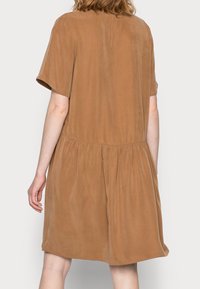 Krátké rukávy, šaty po kolena z lehké hnědé látky s hladkou texturou, s řaseným pasem a volným střihem.