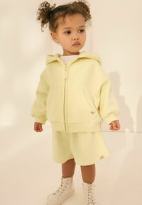 Hoodie zippé jaune clair avec des manches ballon et des poches sur le devant, associé à des shorts assortis. Comprend un petit patch logo et des baskets montantes blanches.