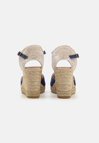 Sandales à semelle compensée avec une tige en beige tissée, des accents en daim bleu marine et un talon enroulé de jute. Comprend une sangle de cheville ajustable et un design à dos ouvert.