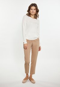 Langärmliges, cremefarbenes Oberteil kombiniert mit beigen, maßgeschneiderten Hosen und hellbraunen Ballerinas; glatte Textur und minimalistisches Design. Die Ohrringe sind kreisförmig.