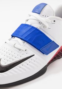 Zapato de entrenamiento Nike blanco con el logo swoosh negro, correa ajustable azul, cordones blancos y suela con tinte rojo visible en el lateral.