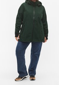 Groene waterdichte jas met capuchon, rits aan de voorkant, zijzakken en zwarte accenten; gecombineerd met blauwe spijkerbroek en sneakers met patroon.