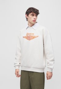 Jeune homme portant un sweat-shirt blanc à col rond avec le logo orange « Dickies Harley-Davidson Motor Cycles » et un pantalon cargo olive.
