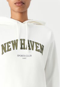 Biela mikina s kapucňou, na prednej strane je grafika s nápisom "NEW HAVEN" v olivovej farbe, nižšie sú v menšom písmene nápisy "SPORTS CLUB" a "GANT".