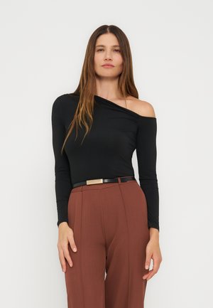 Nő hosszú barna hajjal, fekete egyrészes, hosszú ujjú felsőt és magas derekú barna nadrágot visel, fekete övvel.