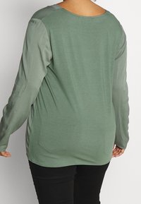 Maglietta verde a maniche lunghe con una texture liscia, scollo rotondo e spacchi laterali, abbinata a pantaloni neri che mostrano una vestibilità comoda.
