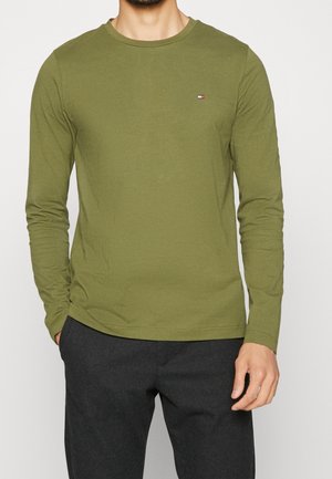 Mann trägt ein olivgrünes Langarmshirt mit einem kleinen Logo auf der Brust und dunkelgraue Hosen, die Hände locker an den Seiten.
