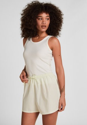 Vrouw met krullend haar, gekleed in een wit mouwloos topje en lichtgele, gestreepte korte broek, poserend met één hand die het trekkoord van de korte broek vasthoudt.