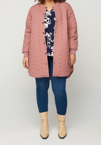 Světle růžový prošívaný kabát s chevronovým vzorem, s patentkami, žebrovanými manžetami, nošený přes námořnické květinové tričko a modré džíny, doplněný o béžové kotníkové boty.