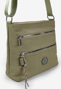 Bolso de hombro de lona verde oliva con dos bolsillos frontales con cremallera, un acento de logo hexagonal y una superficie texturizada. Correa ajustable.