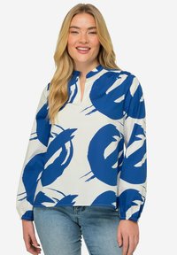 Blusa a maniche lunghe blu e bianca con scollatura a V, caratterizzata da audaci motivi circolari e polsini elasticizzati. Il tessuto appare liscio con una leggera lucentezza.