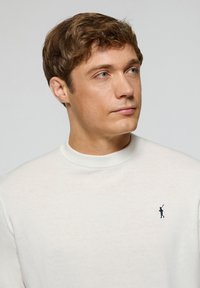 Maglione bianco in maglia con colletto girocollo a coste e un piccolo logo blu scuro sul petto. Tessuto liscio e design aderente.