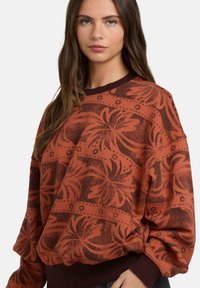 Ceglasty sweter z odważnymi kwiatowymi wzorami, z okrągłym dekoltem i ściągaczami na rękawach. Miękka faktura, luźny krój i długie rękawy.