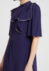 Lila klänning med hög krage, korta flygande ärmar och volangdetaljer. Har metallisk kantning och en flödande, rynkad kjol.