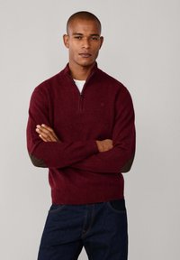 Maglione bordeaux con colletto a zip, caratterizzato da toppe marroni sui gomiti, polsini a costine e una texture morbida. Indossato sopra una t-shirt bianca.